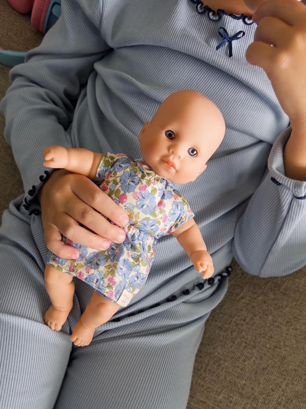 Person holding a baby doll wearing a floral outfit on a carpeted floor.