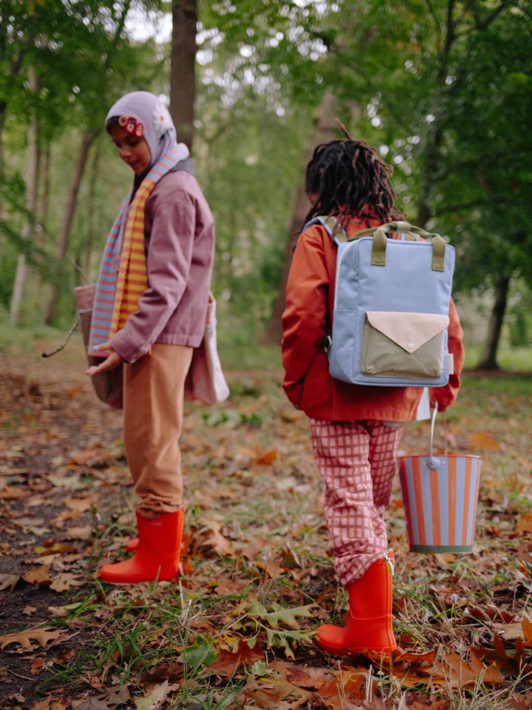 Two people walking in a forest with one carrying a blue backpack and the other holding a striped bag.